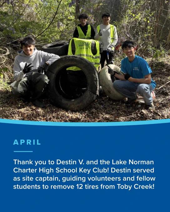 Four high school students pose outdoors with large tires they removed from a wooded area. One student wears a bright yellow safety vest, while the others wear casual clothes and gloves. Sunlight filters through the trees around them.