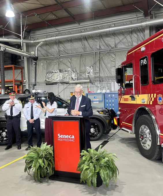 Podium with an individual standing at the podium speaking. The Toronto Fire Chief, Chief of Police and police cruiser and fire truck are in the background.