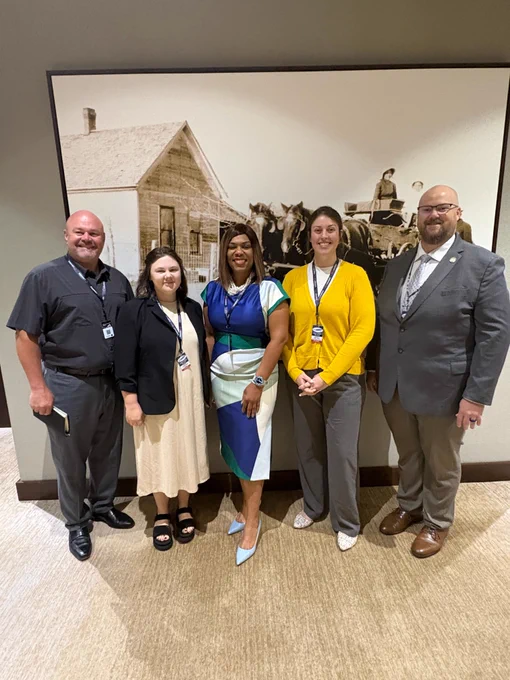 Five professionally dressed individuals stand smiling in front of a large black-and-white photo of a horse-drawn carriage and a rustic house at the Inter-Tribal Emergency Management Coalition's annual conference in Tulsa, OK.