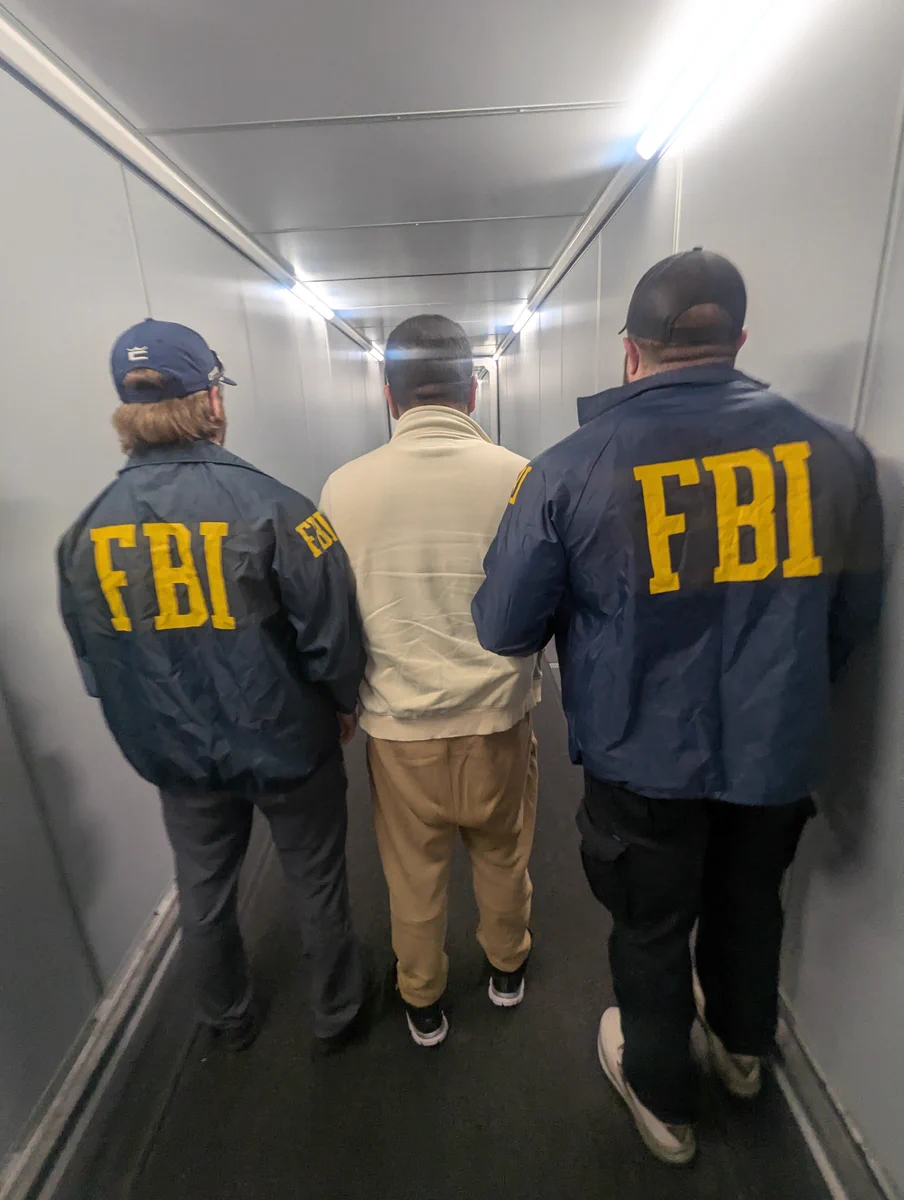 A fugitive extradited from Mexico is photographed with two FBI Special Agents in raid jackets in an airport hallway. 