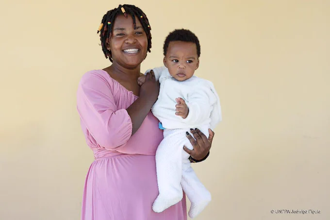 A photo of a woman in a pink dress holding a baby in a white outfit. The mother is smiling broadly.