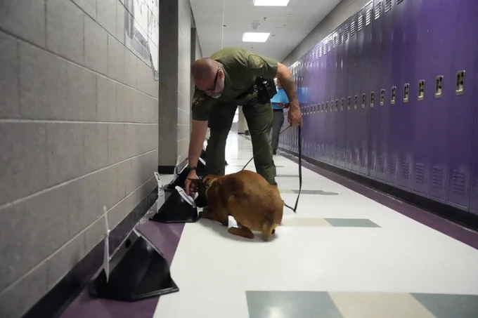 Deputy Austin kneeling in a locker-lined hallway guiding a detection dog as it sniffs scent-training objects placed along the floor.