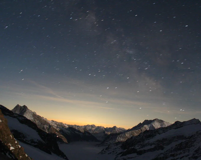 A breathtaking long-exposure shot of a starry night sky above majestic snow-capped mountains. Thousands of streaking stars fill the dark blue sky, with a soft orange glow on the horizon over a vast snowy valley.