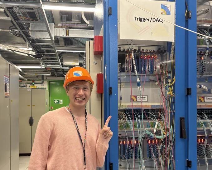 A young man in an orange hard hat and pink sweater smiles, pointing at a blue equipment rack. The rack is filled with glowing electronic components and numerous colorful cables. A sign on the rack reads \"Trigger/DAQ\" with a CMS logo. He is in a facility resembling a data center.