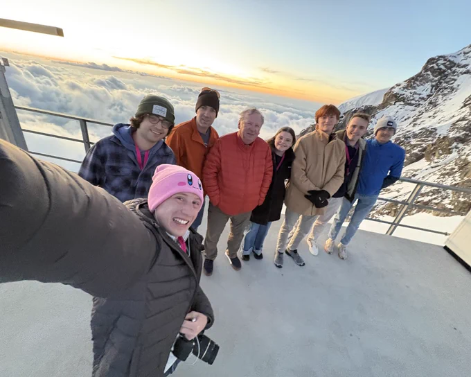 Eight smiling people take a selfie on a mountain observation deck. The person in front wears a pink Kirby beanie and holds a camera. Behind them, a stunning sunset casts an orange glow over a vast sea of clouds, with a snow-capped mountain visible to the right.