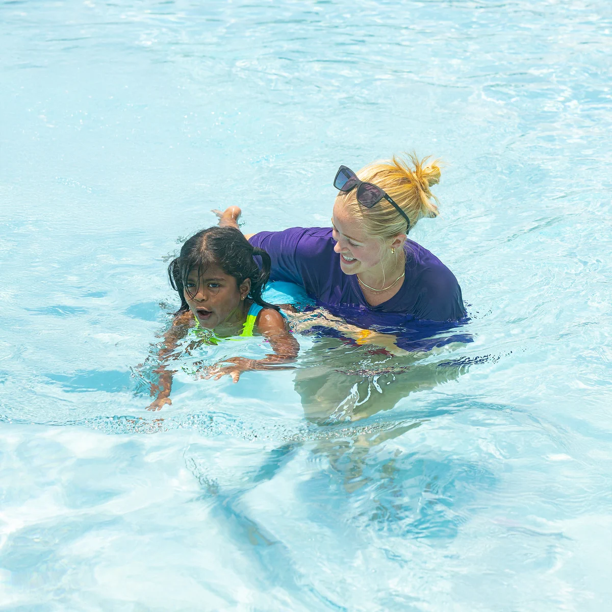 A person in a purple shirt is helping a child learn to swim in a pool. The child is wearing a green swimsuit, and both appear to be focused on the activity in the clear, blue water.