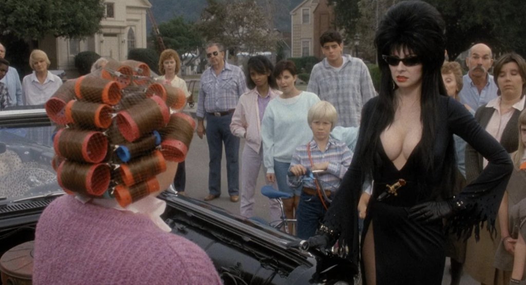 justaida3's tweet photo. 21. ELVIRA: MISTRESS OF THE DARK (James Signorelli, 1988 🇺🇸): Qué maravilla ver por fin en este #BloodySummer a uno de los grandes iconos del fantástico. Una comedia descaradamente gamberra donde la Reina de las Tinieblas demuestra que su mayor poder es un carisma arrollador. 

Todo comienza cuando Elvira hereda una vieja mansión en un pequeño pueblo ultraconservador de Massachussets (ese estado donde, siglos atrás, tuvieron lugar los juicios por brujería de Salem). Lo que parecía la oportunidad perfecta para conseguir dinero para su soñado espectáculo en Las Vegas, se convierte en un choque constante con unos vecinos puritanos incapaces de soportar a una mujer sensual, extravagante y orgullosa de ser diferente.

Me encanta cómo, detrás de los chistes ácidos, ese escote espectacular y el humor picantón, la película es mucho más inteligente de lo que aparenta. Elvira no deja de ser juzgada por cómo viste, cómo habla o cómo se comporta. Las burlas, los insultos e incluso las agresiones que sufre dejan claro que el problema nunca fue ella, sino la intolerancia de quienes la rodean. Pero Elvira nunca intenta cambiar para agradar a nadie. Es ella quien termina poniendo patas arriba un pueblo puritano que parece seguir anclado en el siglo XVII.  

Cassandra Peterson, que ya era todo un fenómeno cultural gracias a Elvira y sus presentaciones de películas de terror en televisión, está absolutamente maravillosa. Su sentido del humor, su capacidad para reírse de sí misma y ese carisma arrollador hacen que resulte imposible no enamorarte de ella. Como tampoco puedes evitar amar a Gonk, su perrito punky cambiaformas

La película destila mucho cariño por el cine de terror clásico: brujas, libros de hechizos, mansiones encantadas, monstruos, serie B, humor absurdo y un espíritu Halloween perfecto. También los guiños a Rambo, Flashdance o Grease son maravillosos.  

No entiendo cómo esta película pudo ser un fracaso en su estreno, pero menos mal que el tiempo ha terminado convirtiéndola en una auténtica película de culto. No será perfecta, pero su protagonista tiene tanto encanto que resulta imposible no rendirse a sus pies.  

🖤 Carisma: sobrenatural
💀 Ranking de pesadilla: not found
🔥 Bloody Summer Approved

#BloodySummer
