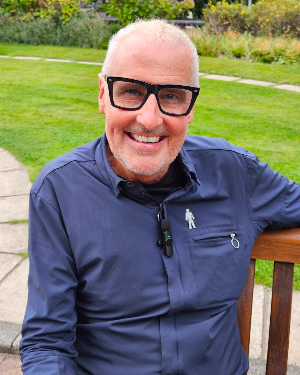 This image shows Dermot Murnaghan sitting on a wooden bench outdoors in a park setting. He is smiling and wearing a dark button-up shirt with a Man of Men pin badge on it. The background includes a grassy area and a path.