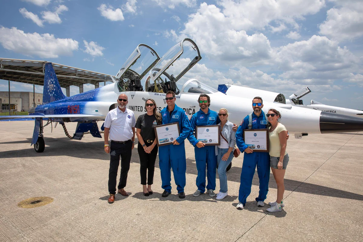 A group of men and women, some wearing blue flight suits and holding awards, stand in front of white and blue fighter jets on a runway.