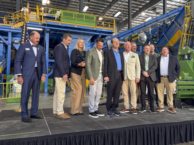 Group of nine professionals standing on a stage inside an industrial facility with large machinery in the background.