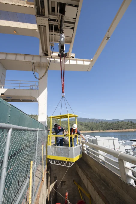 Two workers in safety gear are suspended in a yellow cage lifted by a crane over into a dam.
