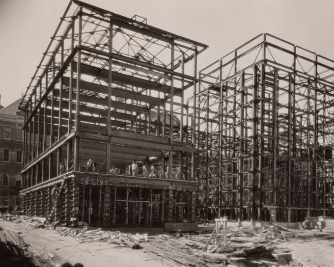 Sepia photo of a bustling construction site with two large, multi-story steel frameworks. The left building has several workers on its lower floors, while the right building shows a complex steel skeletal structure. Debris and wooden planks are scattered across the dusty ground.