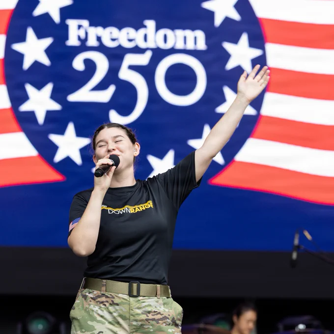 An individual in military uniform sings into a microphone on stage. Behind them, a large screen displays "Freedom 250" with stars and stripes.