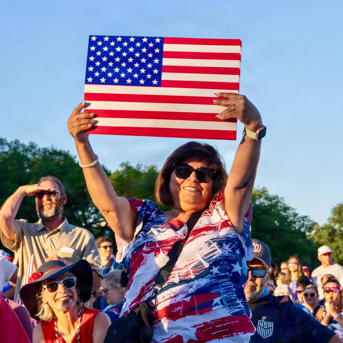 A person in a crowd holds up an American flag with both hands, wearing a patriotic shirt. The scene is outdoors with clear blue skies and trees in the background. Other people in the crowd are seen, with one shading their eyes and another in a USA-themed hat.