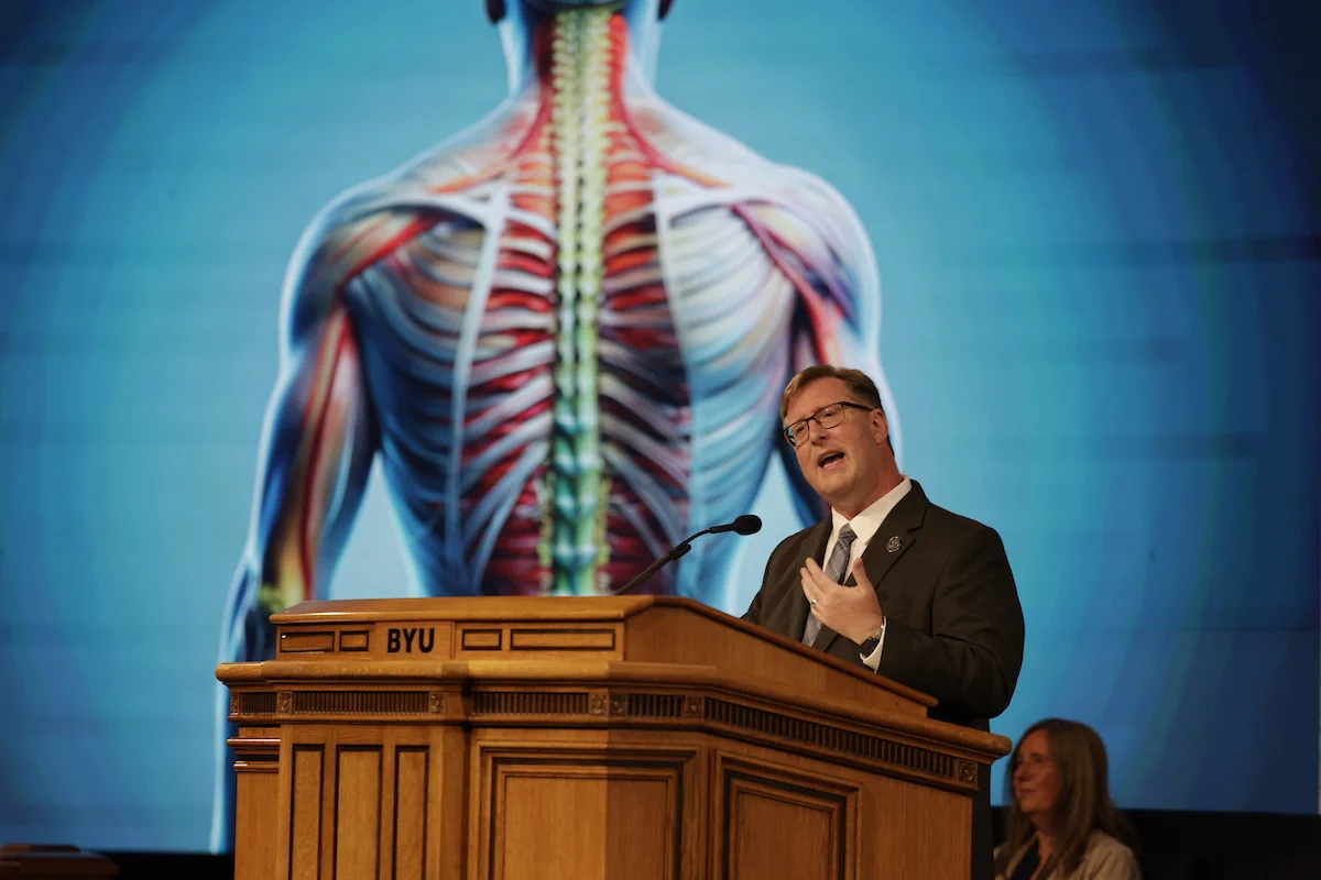 Anton E. Bowden speaks at a podium at BYU Devotional, an image of the human body on the screen behind him