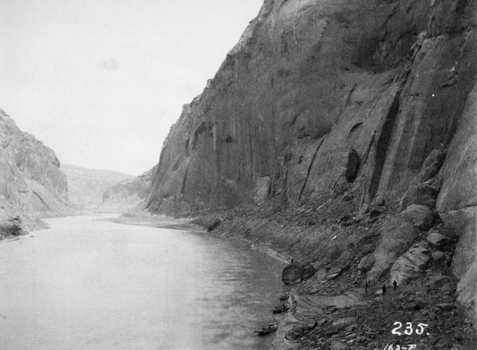 Wide river flowing through steep rocky canyon walls with small boats docked along the shore at the base of the cliffs. 1889 Nims (Stanton Expedition)