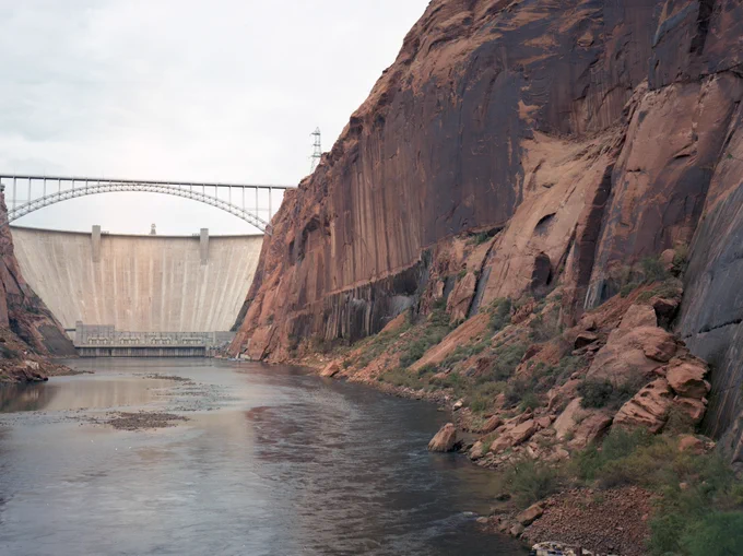 Large concrete dam spanning a river between steep rocky cliffs under a cloudy sky. 1992 USGS.