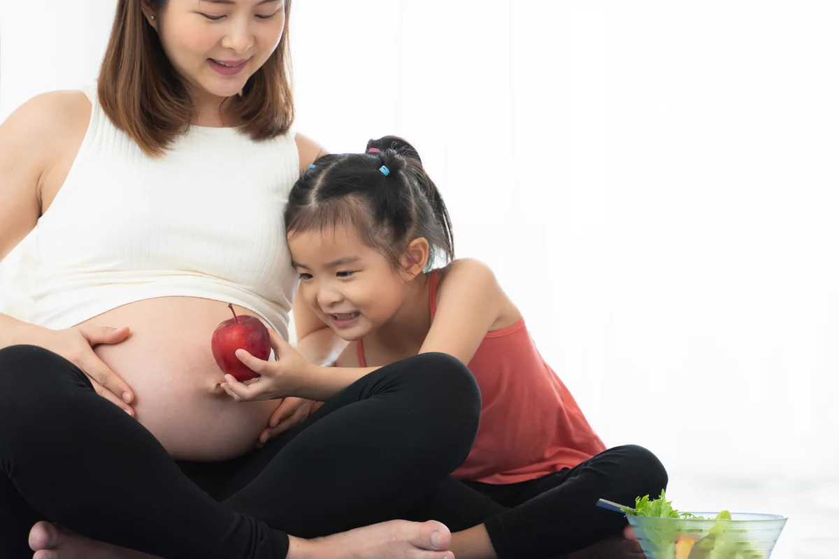 A pregnant mother and her young daughter sitting together, with the daughter holding a red apple against the mother's bump.