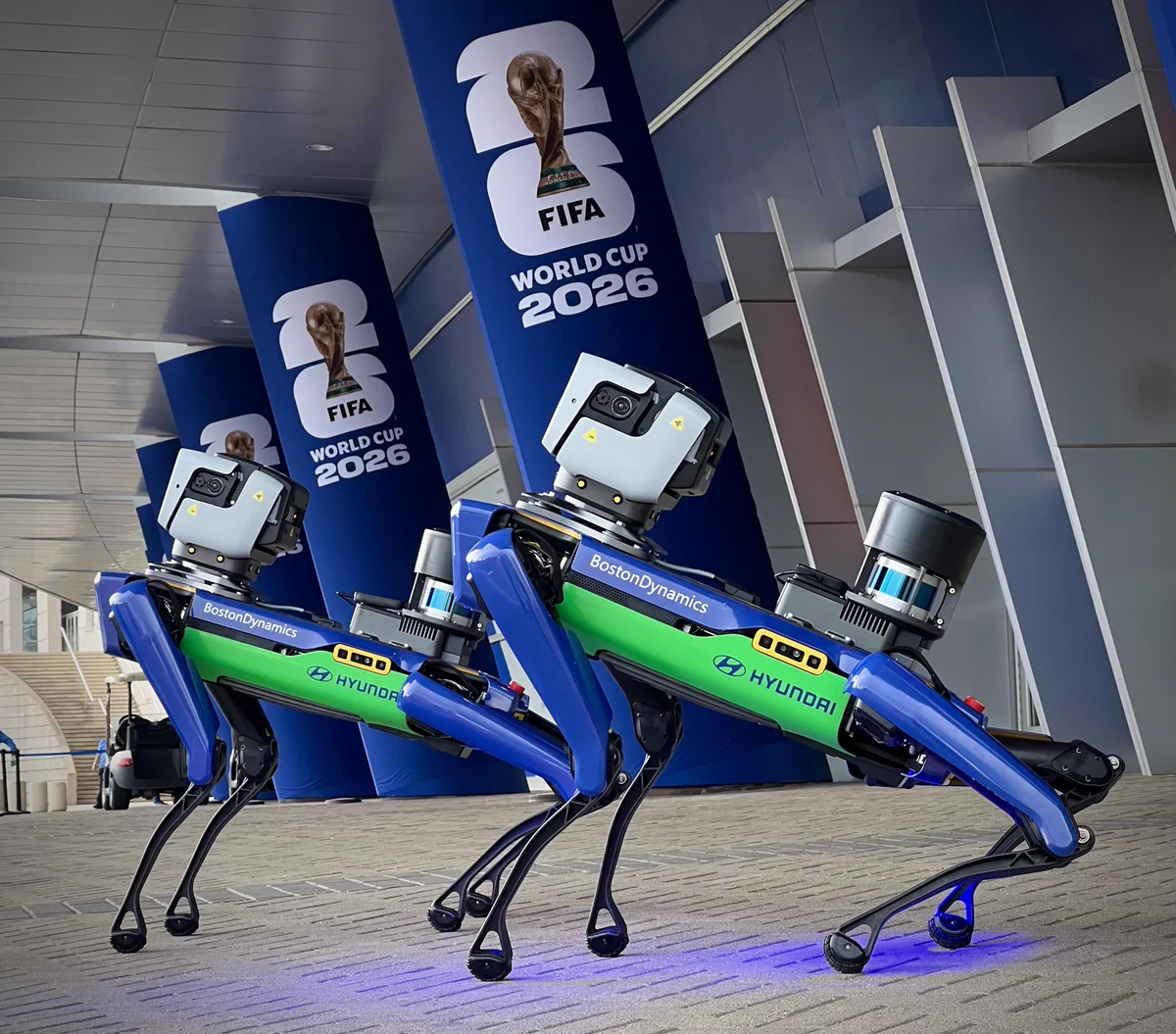 Two Boston Dynamics robotic dogs stand on a paved surface in front of large pillars displaying the FIFA World Cup 2026 logo. The robots have blue and green bodies with visible branding from Boston Dynamics and Hyundai.