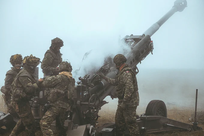 Soldiers in camouflage uniforms operate a large artillery gun emitting smoke during a military exercise.