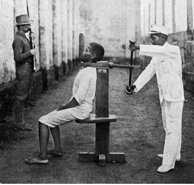 9raTk's tweet photo. This is a picture of a man who is about to be executed using the execution device called the garrote.

The garrote is an execution device that is documented to have been used in the first century BC Rome. This torture method found its way into modern history.

The condemned would sit with a metal clap wrapped around his neck before the executioner turned the screw that would theoretically burst his brainstem, killing him instantly.