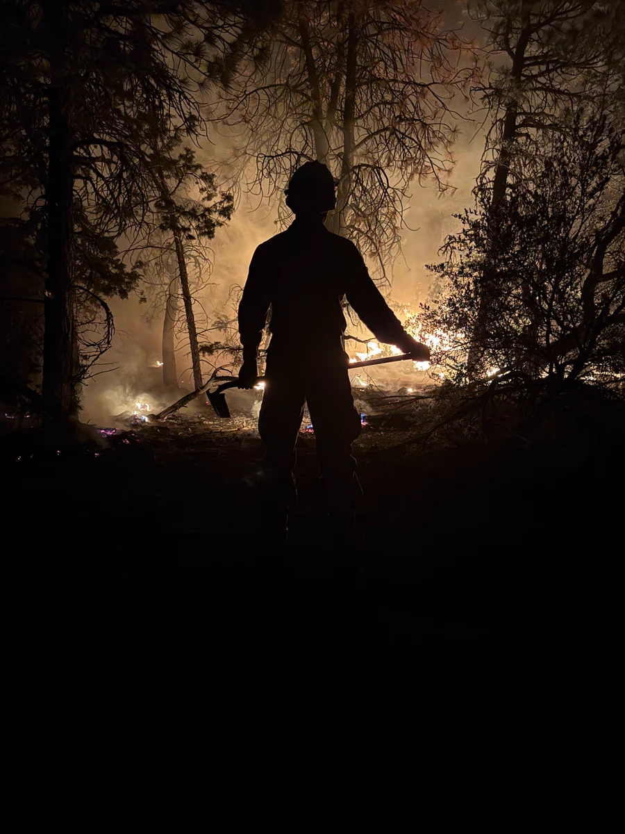 Silhouette of a firefighter holding a tool, standing in front of a blazing forest fire at night.