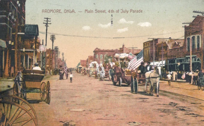 Colorized postcard showing a Fourth of July parade on Main Street in Ardmore, Oklahoma, circa 1911. Horse-drawn wagons and carriages decorated with American flags travel down the wide dirt street while spectators line the sidewalks. Historic brick storefronts and utility poles frame the scene, capturing an early Oklahoma Independence Day celebration.