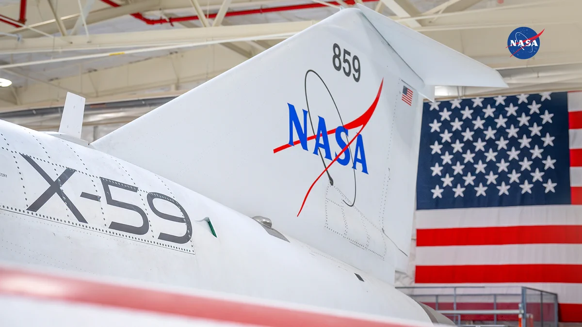 Close-up view of NASA’s X-59 aircraft inside a hangar, showing the tail section with the NASA logo and aircraft number 859. A large American flag hangs prominently in the background, symbolizing U.S. innovation and the future of flight as the nation approaches its 250th anniversary.