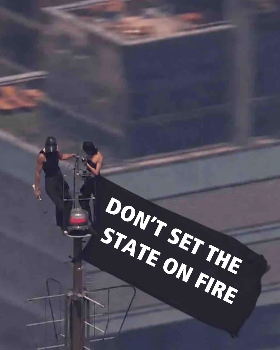 Two individuals who have climbed the spire of the Empire State Building and are flying a black flag that reads "DON'T SET THE STATE ON FIRE"