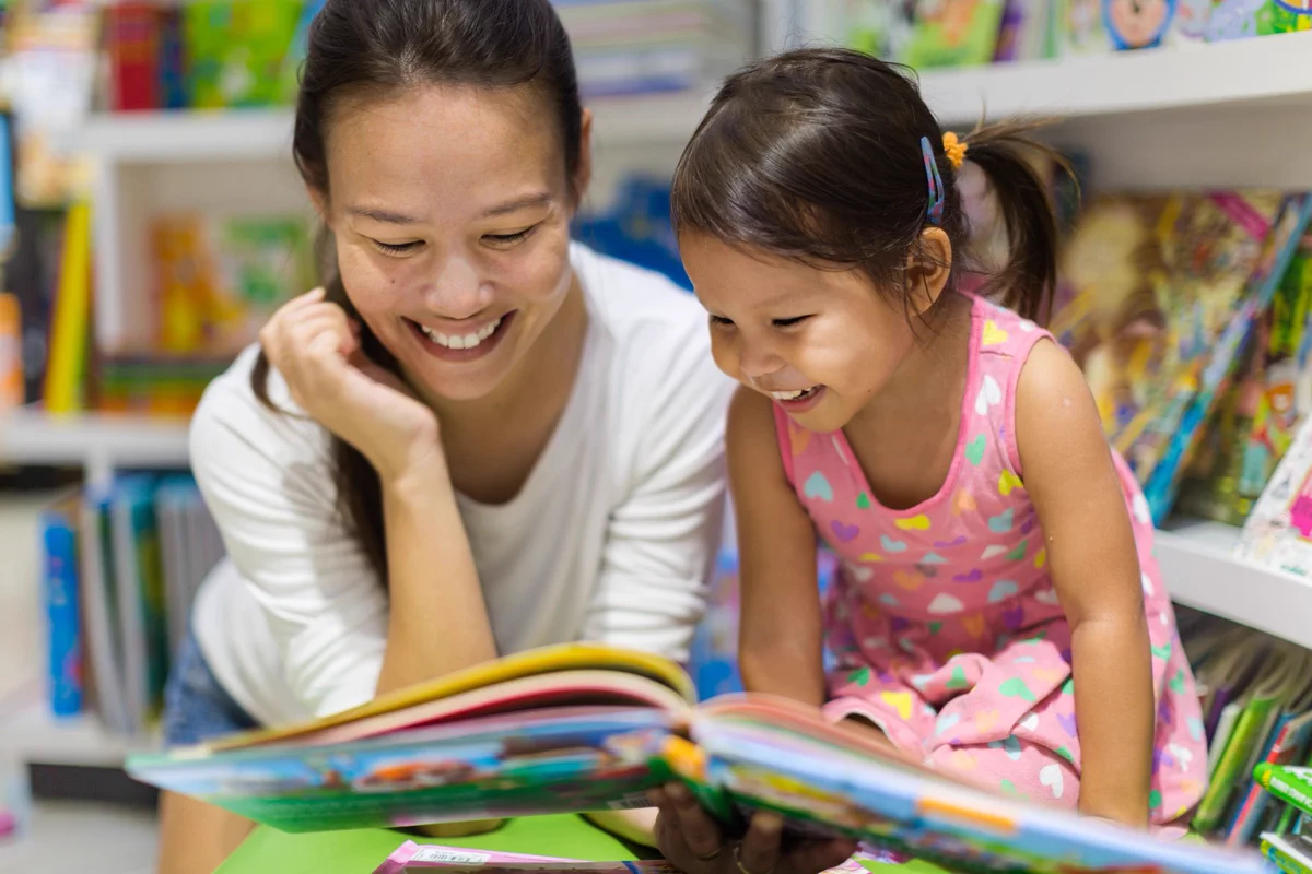 A young child and an adult sit together in a library, looking at an open colourful picture book. Shelves filled with books are visible in the background.