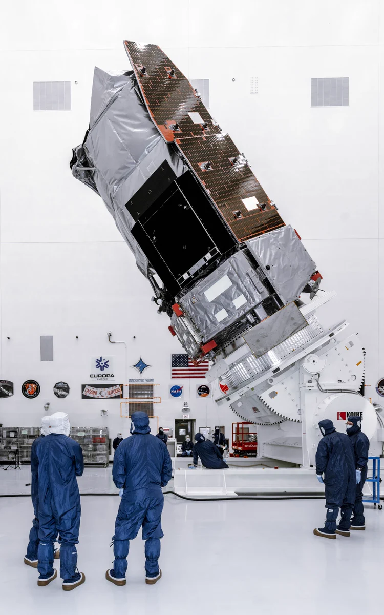 NASA's Nancy Grace Roman Space Telescope, large spacecraft, wrapped in silver thermal covering is tilted upright on a massive white rotation stand inside a clean room at NASA's Kennedy Space Center in Florida on Thursday, June 25, 2026. Several technicians in blue clean-room suits and protective gear stand around the base, observing the operation. The spacecraft’s solar arrays are partially deployed overhead. Mission logos, an American flag, and clean-room equipment line the white walls in the background. Photo credit: NASA/Sydney Rohde (Rocz)
