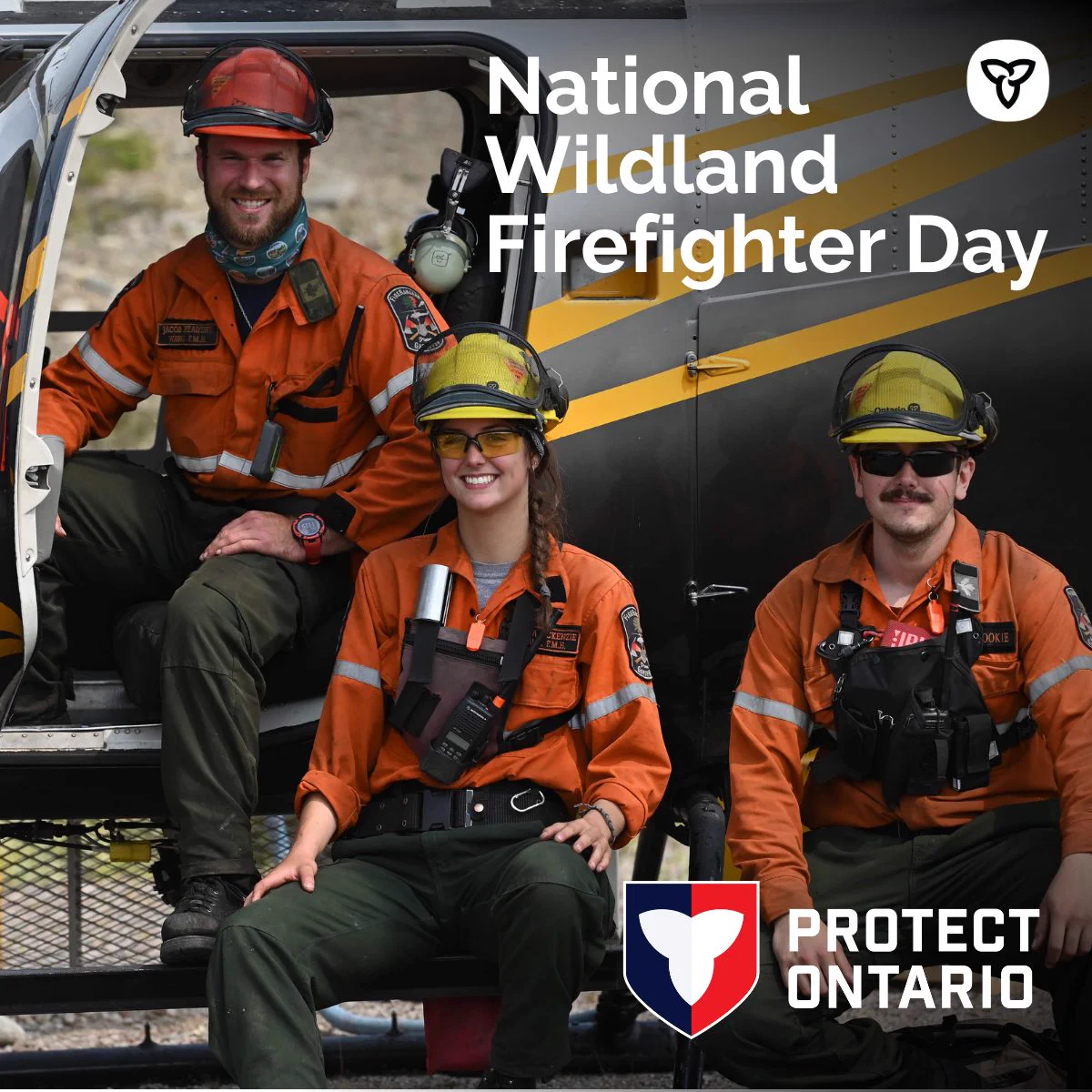 Three wildfire firefighters in orange uniforms and helmets pose in front of a helicopter, ready for action in a rugged outdoor setting.