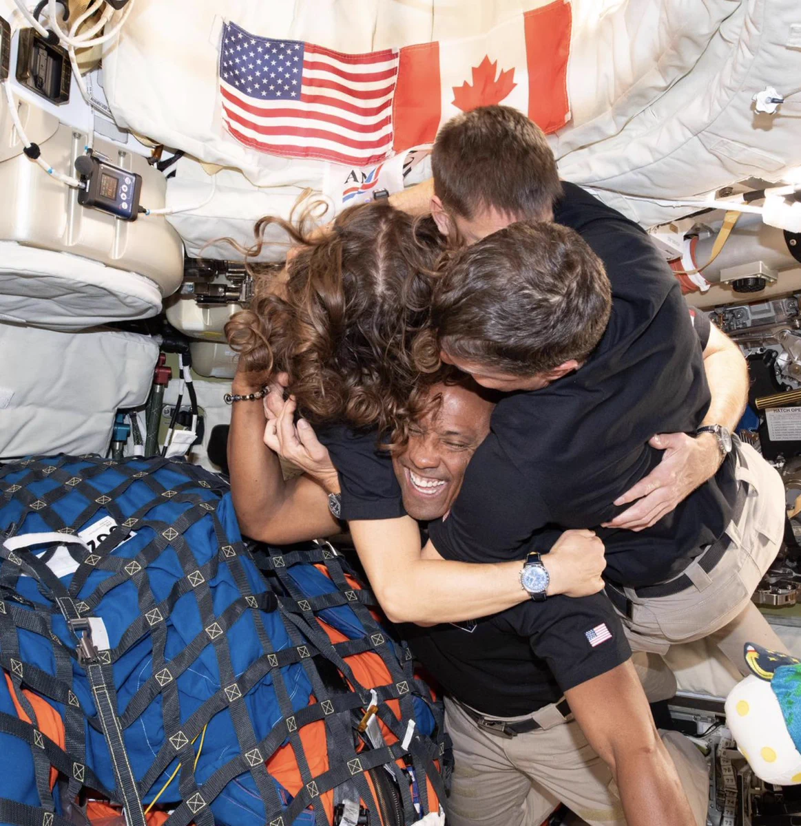 Four astronauts, floating in microgravity inside a capsule, embrace beneath two small flags, from the U.S. and Canada. 