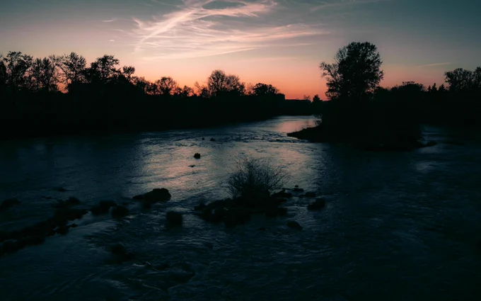 A tranquil river flows beneath a dusk sky, with the silhouette of trees along the water's edge. The sky features streaks of clouds and warm colors transitioning to night.