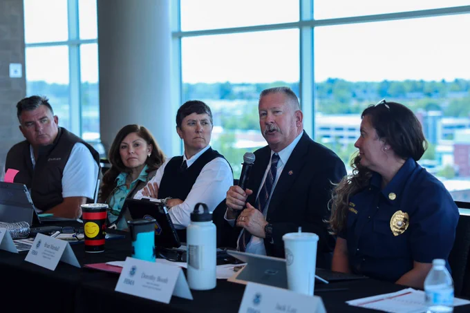 Image shows a group of people sitting at a table and a man in a suit talking into a microphone. 