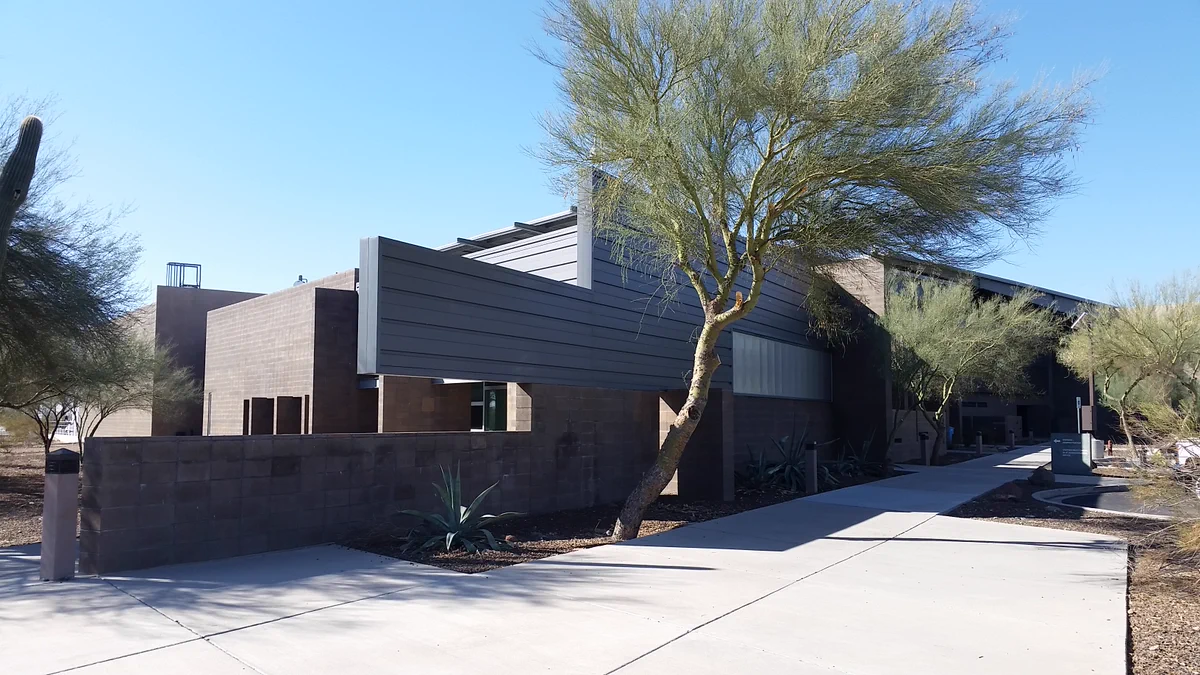 An exterior shot of the North Gateway Transfer Station. It's a gray, modern industrial style buildilng with palo verde trees in front. The sky is clear and blue during daytime.