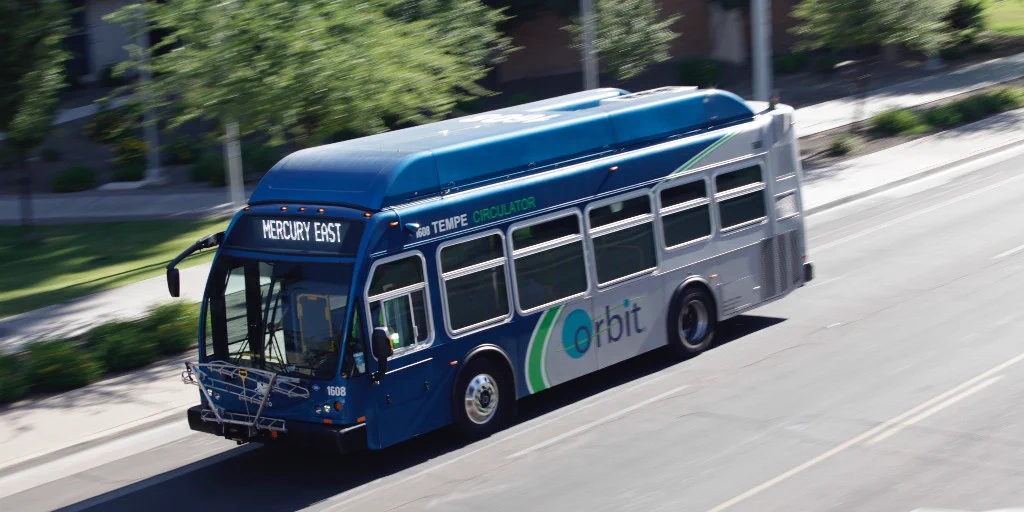 A blue and white public bus labeled "Tempe Orbit Mercury East" travels along a road. Trees and a building are in the background.