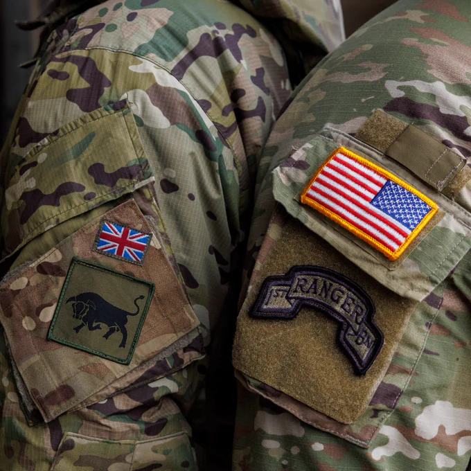 Two people wearing military uniforms are shown from the back. One uniform features a patch with the Union Jack and a black bull silhouette. The other uniform displays an American flag patch and a "Ranger" patch. Both uniforms are in camouflage pattern.
