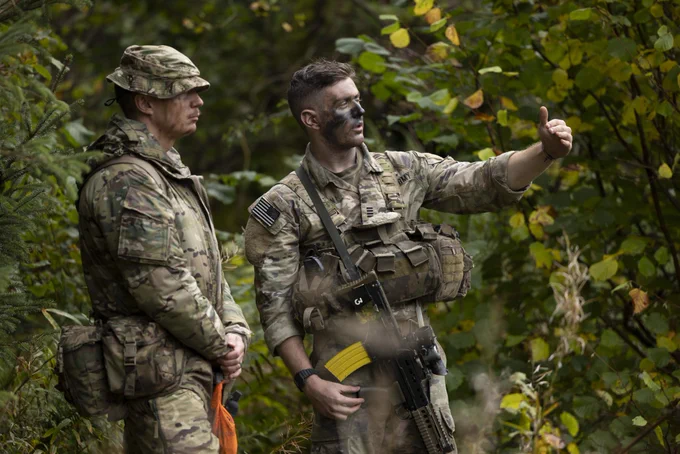 Two individuals in military camouflage uniforms are in a forested area. One is pointing towards something off-camera while the other listens intently. The scene is surrounded by dense green foliage, creating a natural backdrop.