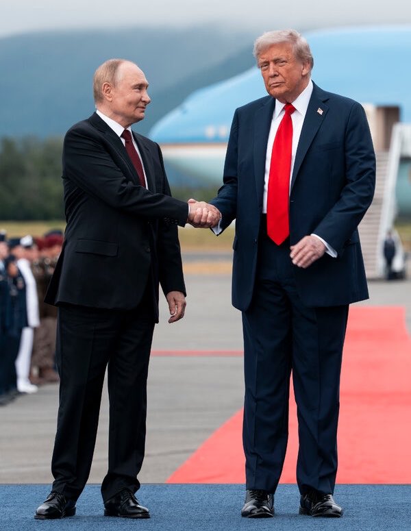 Putin and Trump shaking hands on a red carpet with an aircraft and honor guard behind them