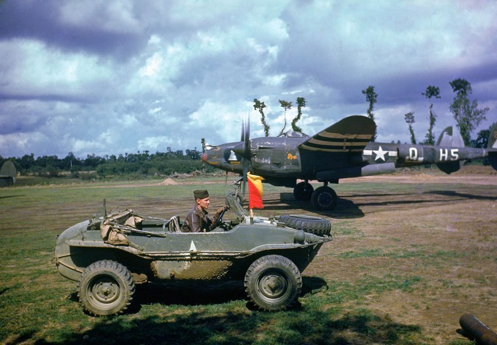 VoicesofWW2's tweet photo. A P-38 fighter plane sat in the background as the pilot arrived in a captured German vehicle, France, 1944. https://t.co/8QpXw0B05F