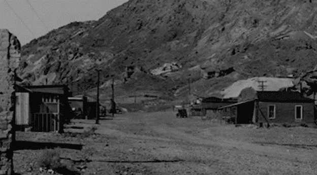 A black-and-white photo of a deserted town with a dirt road running through it. There are small wooden buildings on either side, set against a backdrop of rugged, barren hills. 