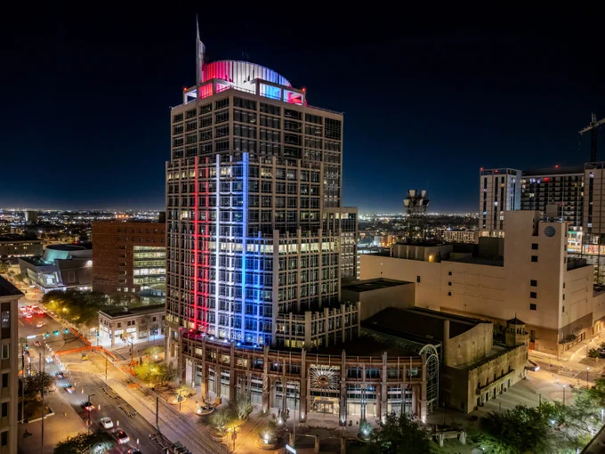 Phoenix City Hall is illuminated with red, white, and blue lights against the night sky. Surrounding buildings and city streets are visible, creating a vibrant cityscape.