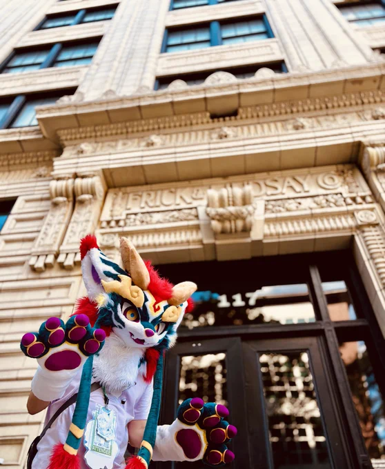 A person wearing a vibrant, colorful fursuit with horns and large paws stands in front of the entrance to The Andy Warhol Museum. The building features ornate architectural details and a large entrance.
