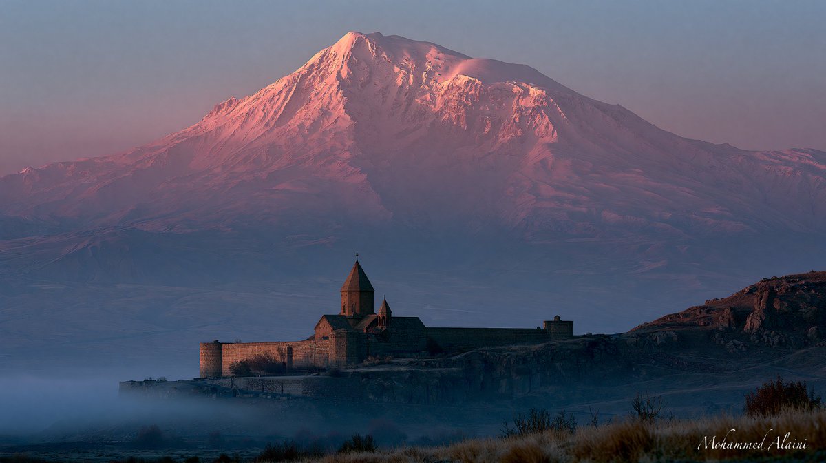 Diaspra's tweet photo. صبا الخير لكم من خور فيراب، ارمينيا

الدير ليس كبيراً. كنيسة حجرية بسيطة. جدران عتيقة. قبة مدببة. لكن ما خلفه هو الذي يصنع المشهد. جبل أرارات. خمسة آلاف ومئة وسبعة وثلاثون متراً من الثلج والصخر يرتفع في السماء كأنه ليس من هذه الأرض. قمة تغطيها الثلوج الدائمة. تقف خلف الدير صامتة. شاهدة. أبدية.

خور فيراب تعني "الحفرة العميقة" بالأرمنية. وليس اسماً. إنه وصف. هنا، في القرن الثالث الميلادي، ألقى الملك تيريداتس الثالث برجل اسمه غريغوري في حفرة مظلمة تحت الأرض. ثلاثة عشر عاماً. في الظلام. في العزلة. في الحفرة التي لا يصلها ضوء. كان غريغوري مبشراً مسيحياً في مملكة لم تعرف المسيحية بعد. قالوا له: توقف عن هذا. رفض. فألقوه في الحفرة. لكن غريغوري لم يمت.

بعد ثلاثة عشر عاماً، مرض الملك. جن جنونه. لم يشفه طبيب. تذكر أحدهم السجين في الحفرة. أخرجوه. صلى غريغوري. شفي الملك. وحدث ما لم يحدث في أي بلد من قبل: اعتنقت أرمينيا المسيحية. أول دولة في التاريخ تتخذ المسيحية ديناً رسمياً. سنة ٣٠١ ميلادية. قبل روما. قبل بيزنطة. قبل العالم.

بُني الدير فوق الحفرة. الكنيسة الصغيرة التي تراها اليوم تعود إلى القرن السابع. لكن الأرض تحتها أعمق. هناك سلالم حجرية تنزل إلى بئر. إلى قاع الحفرة حيث قضى القديس سنواته. والحجاج ينزلون. واحداً بعد واحد. يلمسون الجدران. يضيئون الشموع.

وخارج الدير، الجبل. أرارات. الذي حمله الأرمن في دمهم. والذي راح للأتراك في معاهدة. لكنه لا يزال ينتمي إلى الأرمن بالروح. يقف هناك. على الحدود. ثلجه لا يذوب. والكنيسة عند قدميه تصلي. منذ سبعة عشر قرناً. وما زالت.
