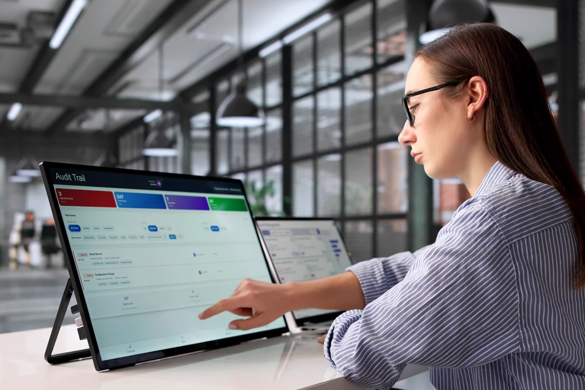 A person is using a touch-screen monitor displaying a software named "Audit Trail" in a modern office setting. The workspace has large windows and pendant lights. Another monitor is visible in the background.