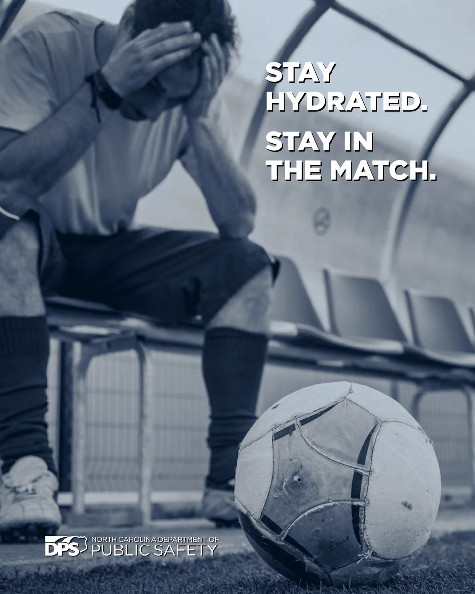 A person sits on a bench in a soccer stadium, head resting in hands. A soccer ball is on the ground nearby. Text on image reads, "Stay hydrated. Stay in the match." The North Carolina Department of Public Safety logo is visible.