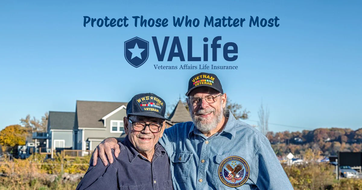 Two veterans wearing military hats stand arm in arm outdoors with houses and trees in the background under a clear sky.
