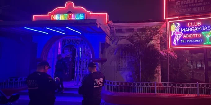 The image shows a nightclub with bright neon signs reading "Night Club" and "Club Margaritas Abierto." Several individuals in uniform are standing outside the entrance, illuminated by pink and blue lights.