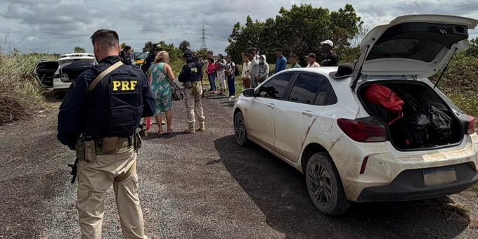 A group of people is gathered at a roadside check, led by an officer with "PRF" on the back of his uniform. The trunk of a white car is open and filled with bags.